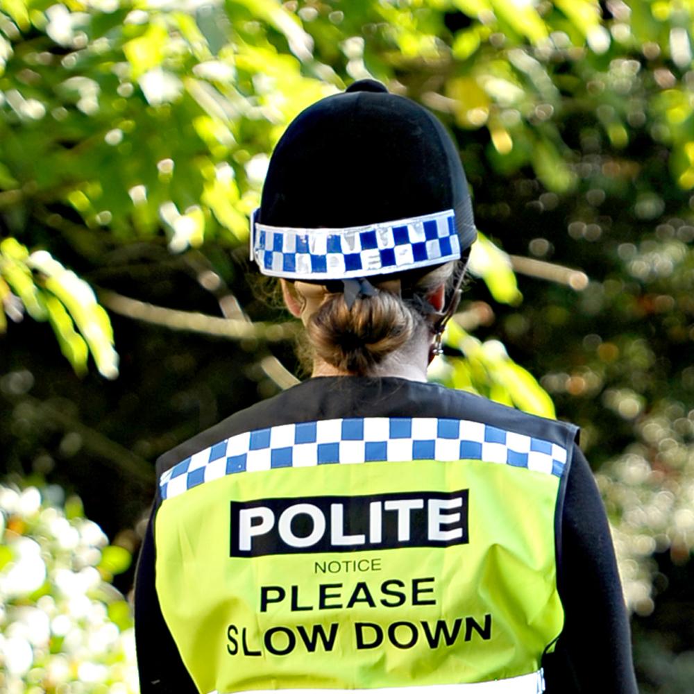 Police officer wearing Equisafety Polite Hat Band holds Please Slow Down sign outdoors.