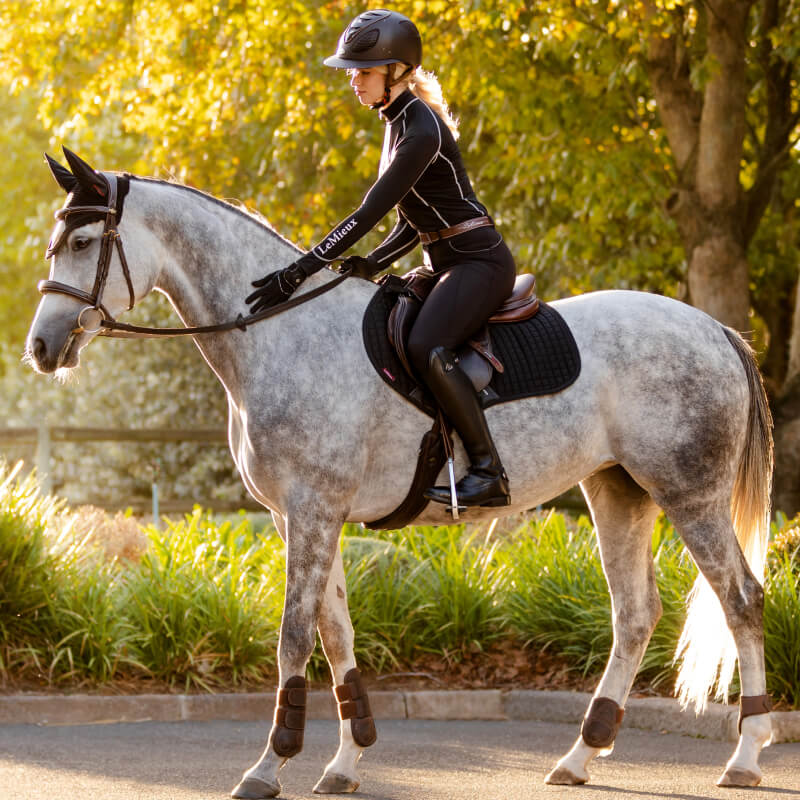 Rider on gray horse uses LeMieux Suede Close Contact Square Black pad near green trees.
