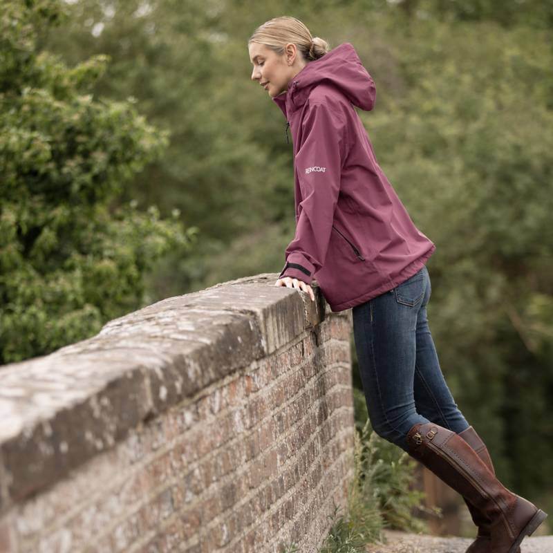 Woman in Reinwear Reincoat Smock Lite Adult Bloom leans on stone wall, trees behind her.