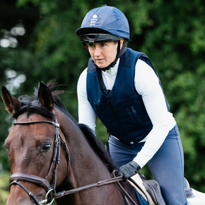 A focused rider in a helmet wears the Racesafe Motion Air Jacket Navy Adult on a dark brown horse.