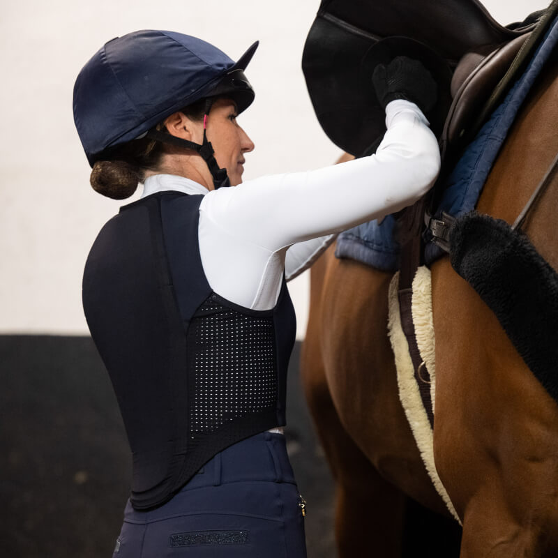Rider in Racesafe Motion Lite Adult Back Protector adjusts a saddle on a brown horse indoors.
