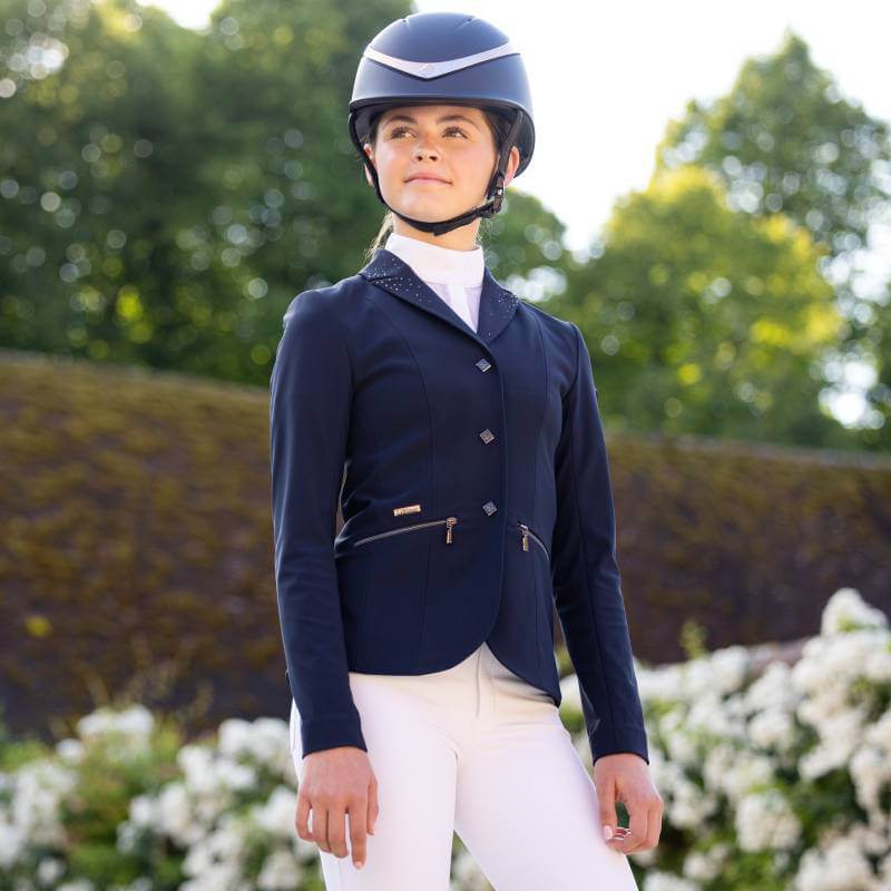 Wearing a LeMieux Junior Pro Show Jacket Navy, a rider stands outdoors among greenery and flowers.