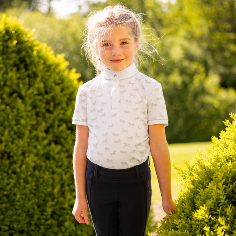 Smiling girl outdoors in a LeMieux Mini Bella Show Shirt White between green bushes.
