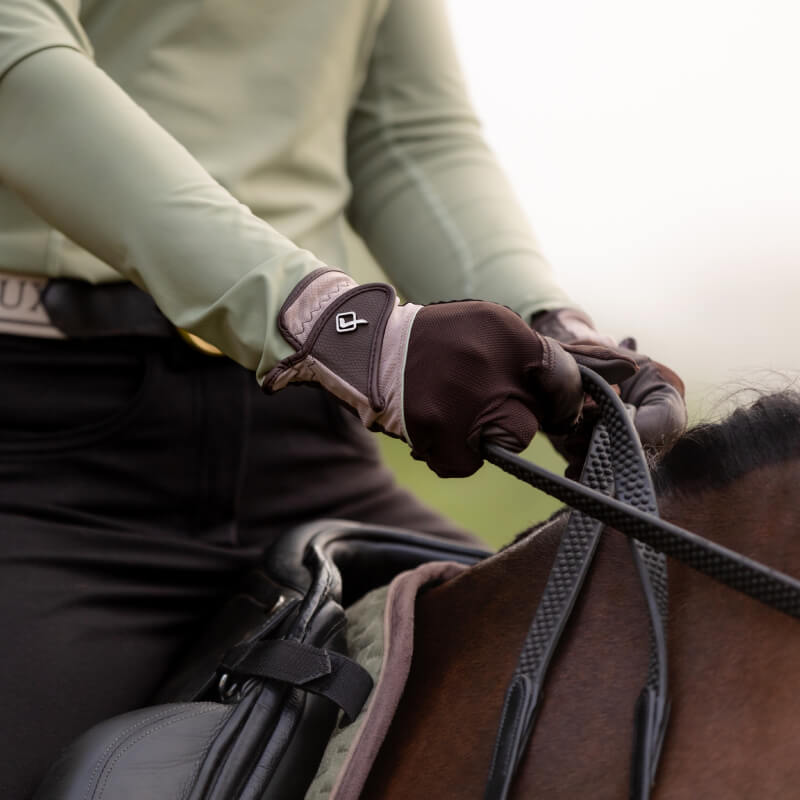 Close-up of LeMieux Pro Mesh Glove Fern/Brown gripping horse reins, saddle partly visible.