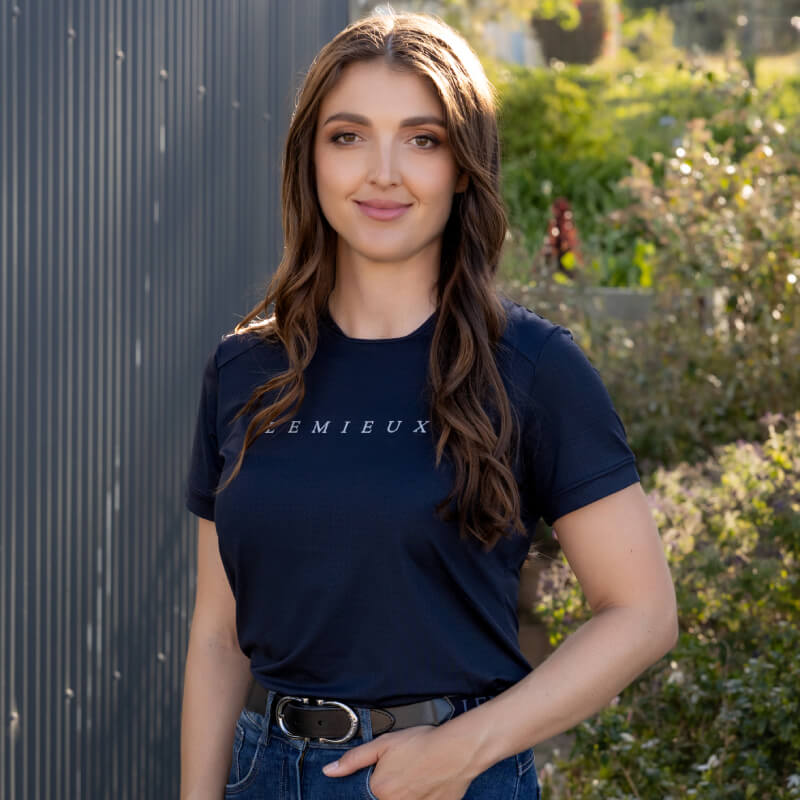 Woman with long brown hair wears a LeMieux Sports T-Shirt Navy by a metal fence and greenery.