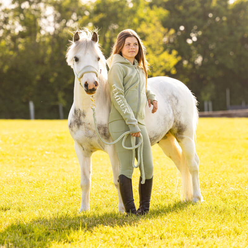 Woman in a LeMieux Young Rider Heidi Hoodie Fern stands by a white horse on a grassy field.
