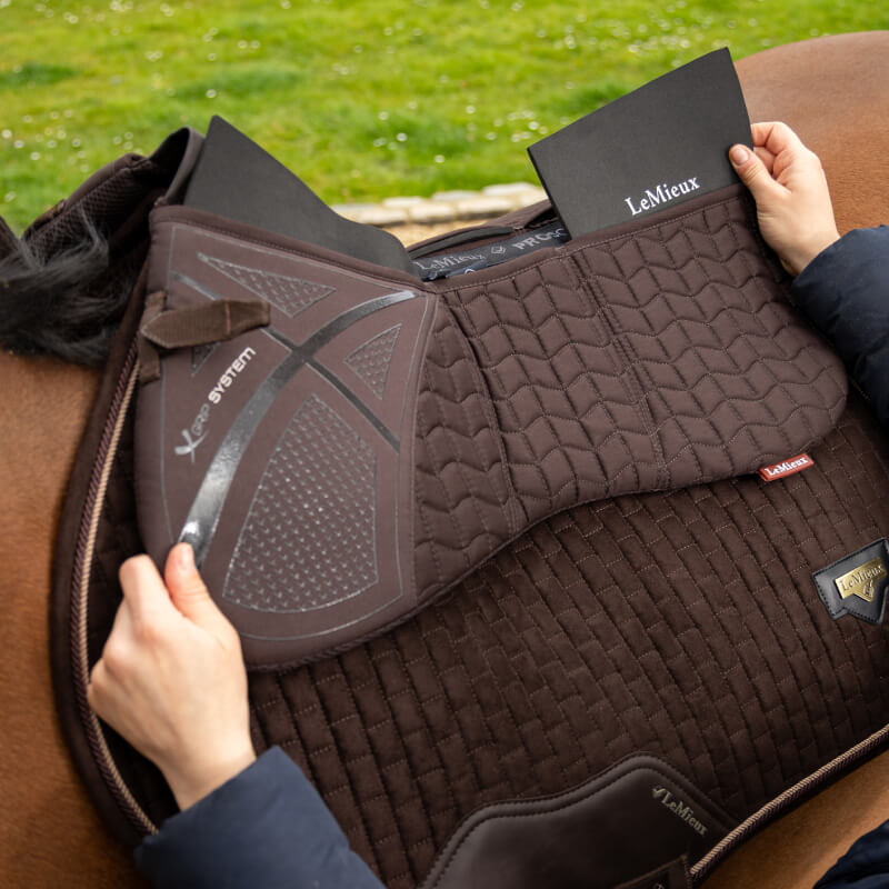 Hands placing a brown LeMieux ProSorb Plain 3 Pocket Half Pad on a horse, grass in background.
