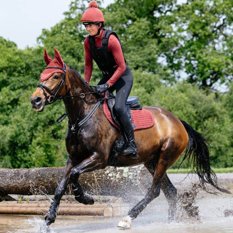 Rider in red on brown horse crosses water with a LeMieux Suede Close Contact Square Russet.