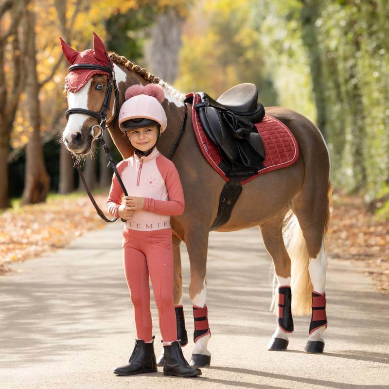 Child in pink stands by pony in LeMieux Suede Close Contact Square Russet on a tree-lined path.
