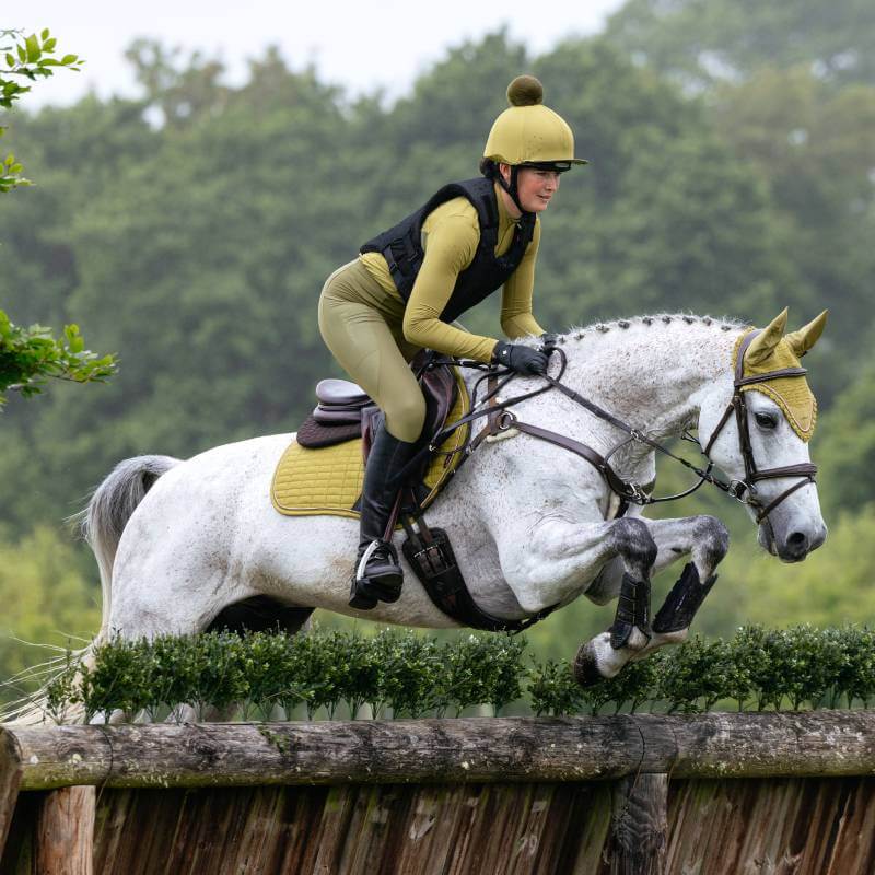 Rider in yellow gear jumps white horse with LeMieux Suede Close Contact Square in Gooseberry.
