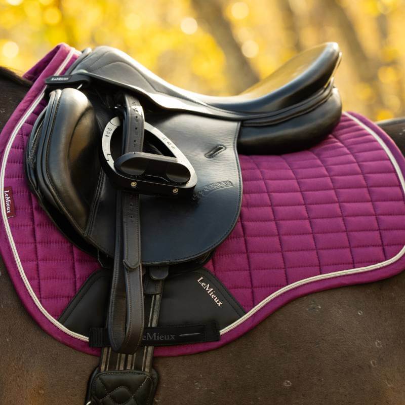 A black saddle on a LeMieux Suede GP Square Blackberry, horse and blurred trees in the background.