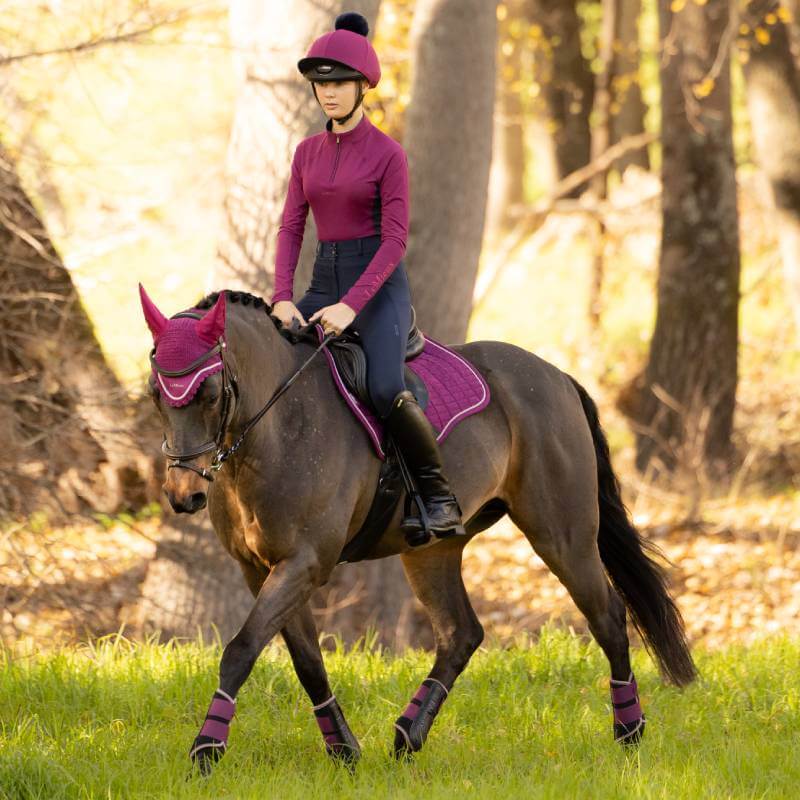 Person in maroon riding a brown horse with a LeMieux Suede GP Square Blackberry in sunlit forest.