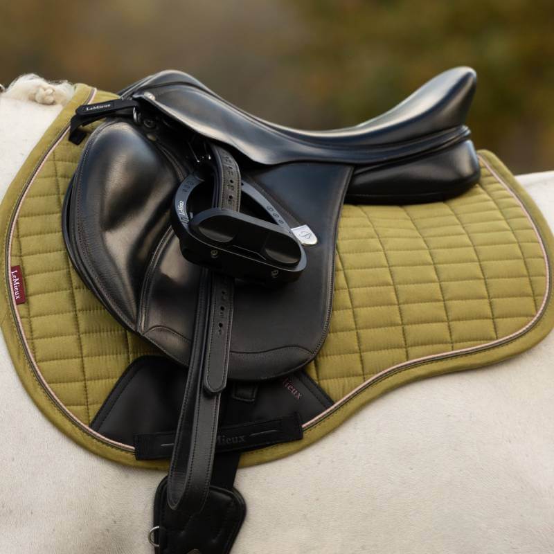 A black leather saddle on a LeMieux Suede GP Square Gooseberry pad on a white horse.
