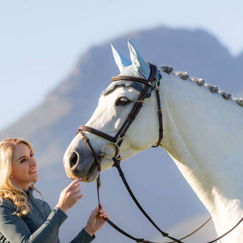 Woman smiling at a white horse in a LeMieux Loire Fly Hood Petrol, mountains in background.