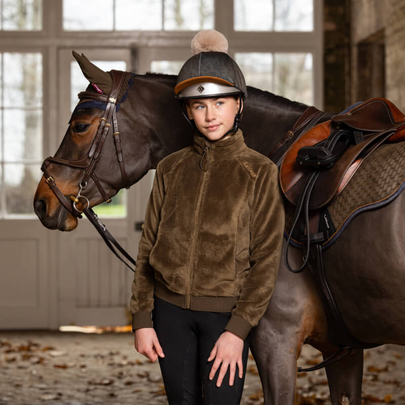 Young rider in a LeMieux Young Rider Libby Fleece Alpine stands by a saddled horse indoors.