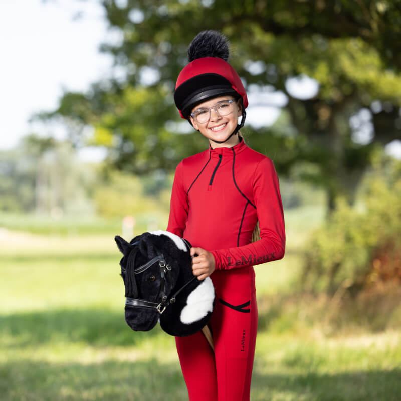 Smiling child in red holds a LeMieux Hobby Horse Razzle on a grassy outdoor field.