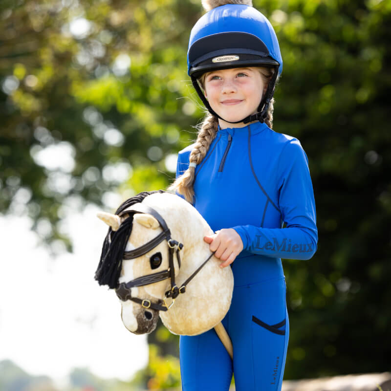 Girl in blue LeMieux outfit and helmet holds a LeMieux Hobby Horse Dream outdoors.