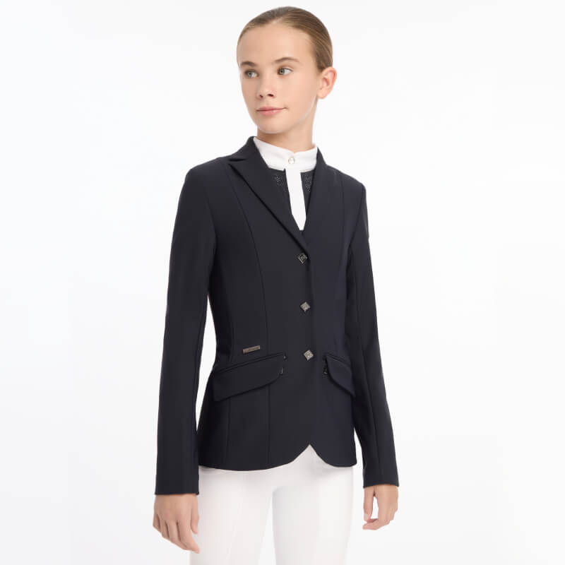A girl wears a LeMieux Young Rider Kennedy Show Jacket Navy and white pants against a white background.