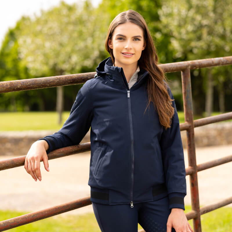 Woman wears LeMieux Ladies Team Jacket Navy by a metal fence with trees in the background.