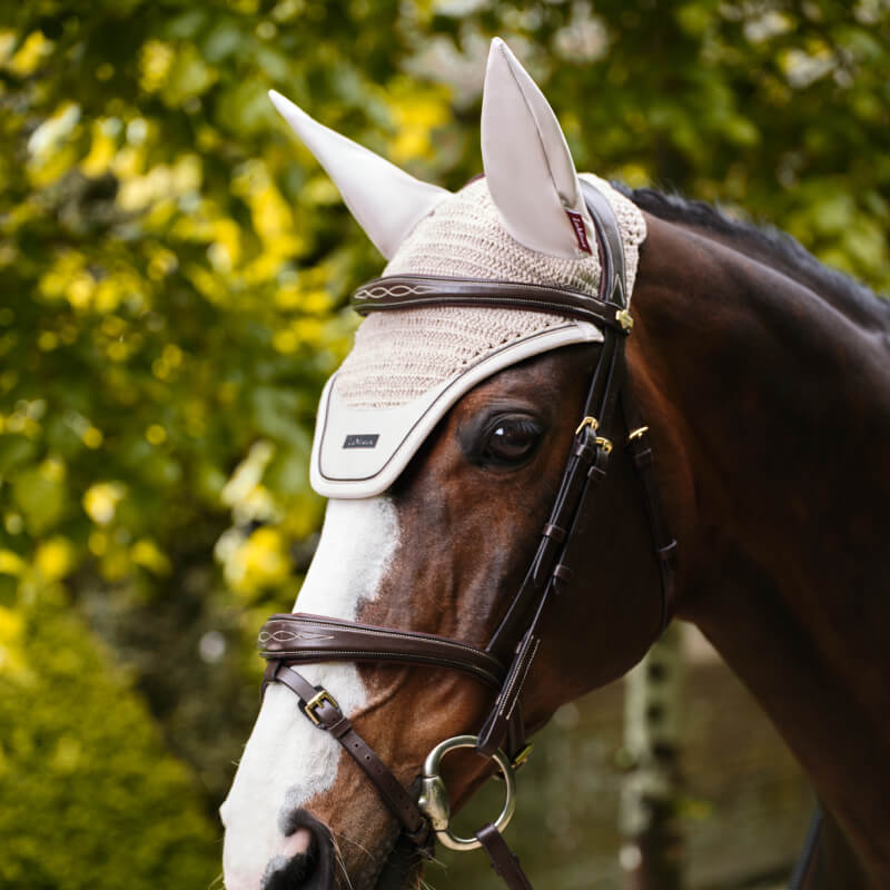 LeMieux Essence Acoustic Fly Hood Stone keeps the brown horse calm outdoors amid green foliage.