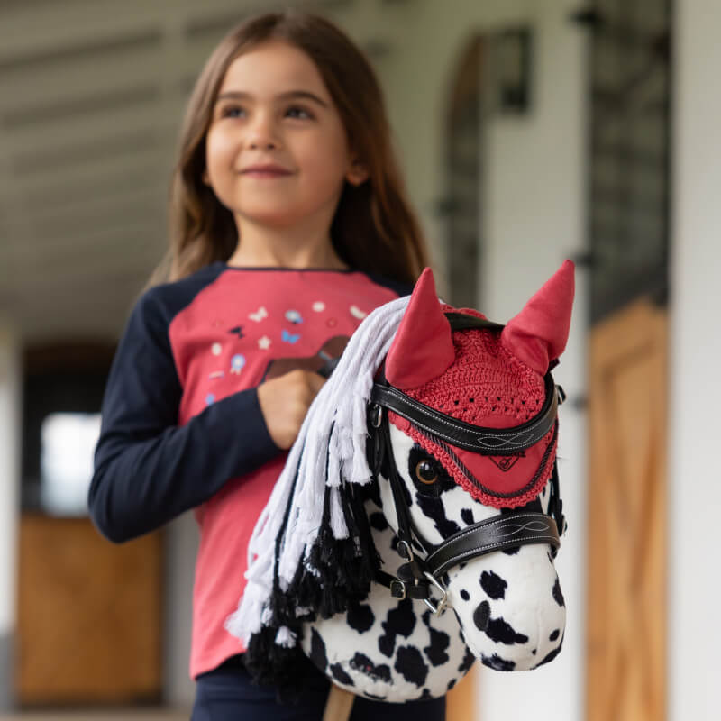 Smiling girl holding a LeMieux Hobby Horse Dakota with red ear bonnet and bridle.