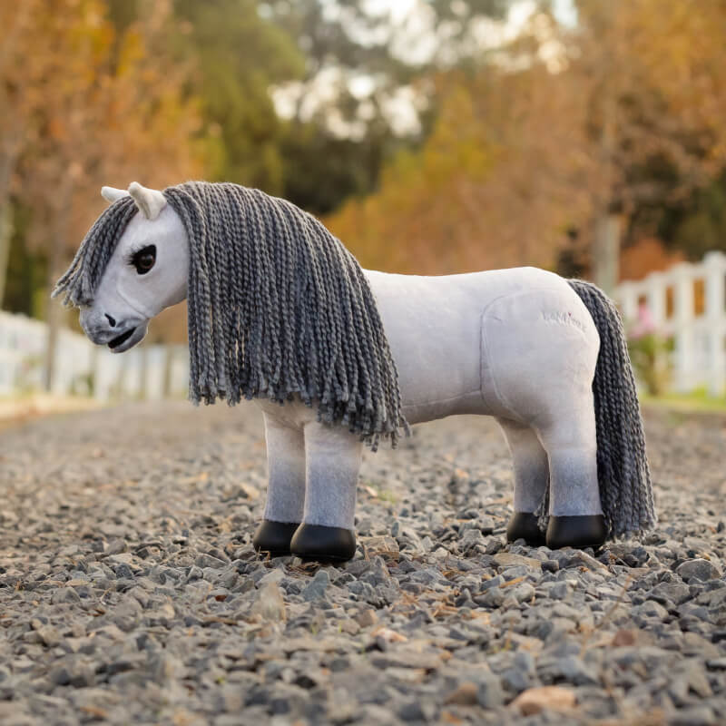 LeMieux Toy Pony Blanca with a gray mane stands outdoors on a gravel path.