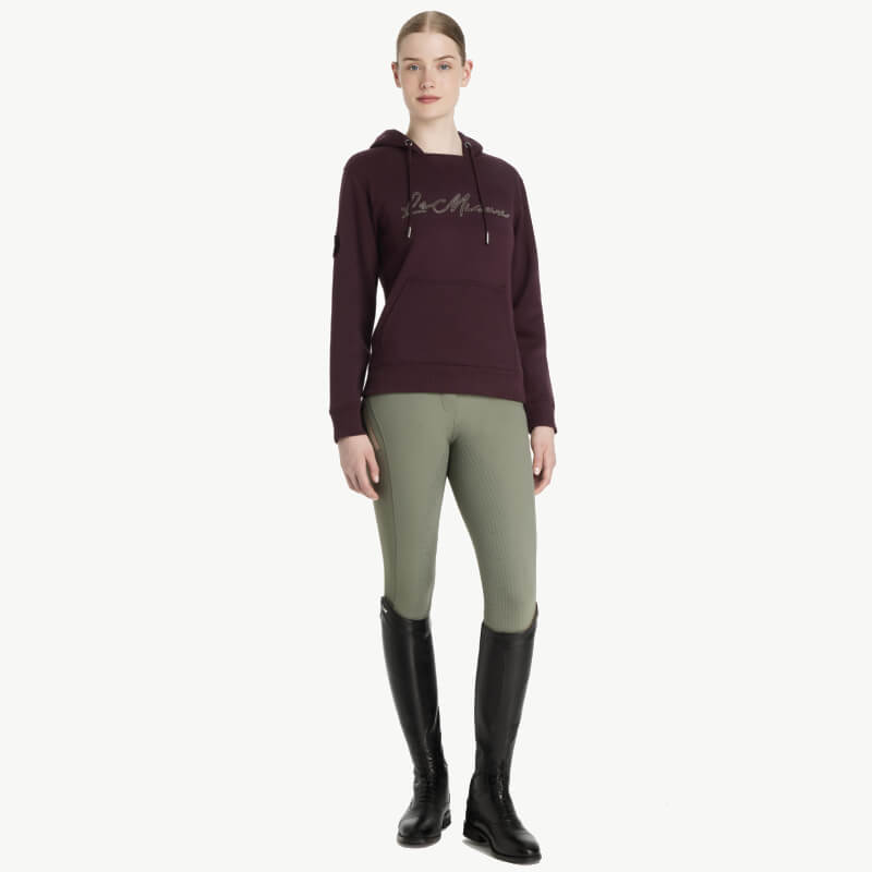 Woman in a LeMieux Elspeth Hoodie Damson, green riding pants, and black knee-high boots.
