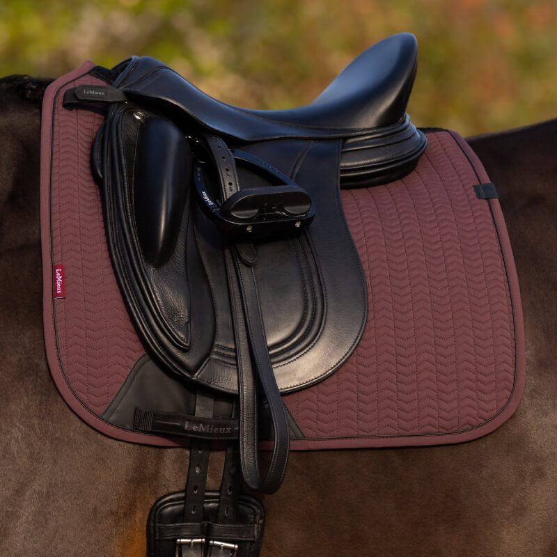 Black saddle with LeMieux Essence Dressage Square Damson pad, outdoors.