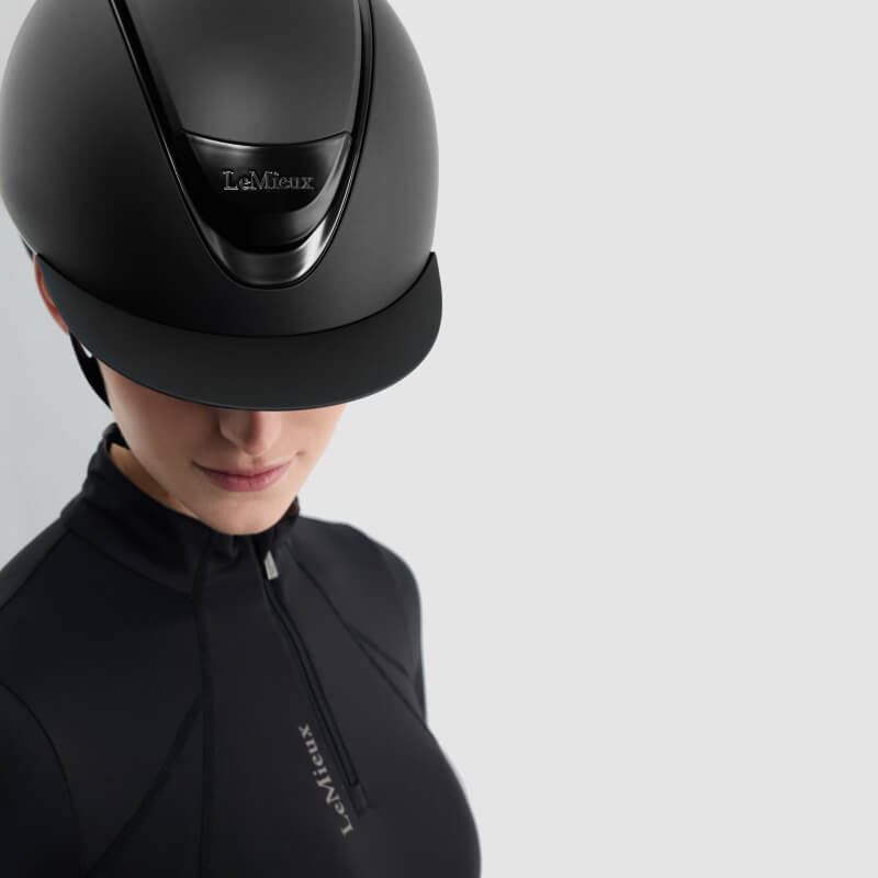 Person wears a LeMieux Verv® Matte Black Competition Helmet and LeMieux top, plain background.