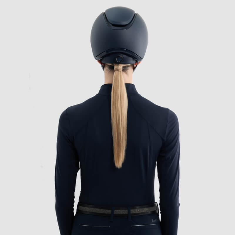 Woman with long blonde ponytail in LeMieux Verv® Matte Navy helmet and dark jacket, seen from behind.
