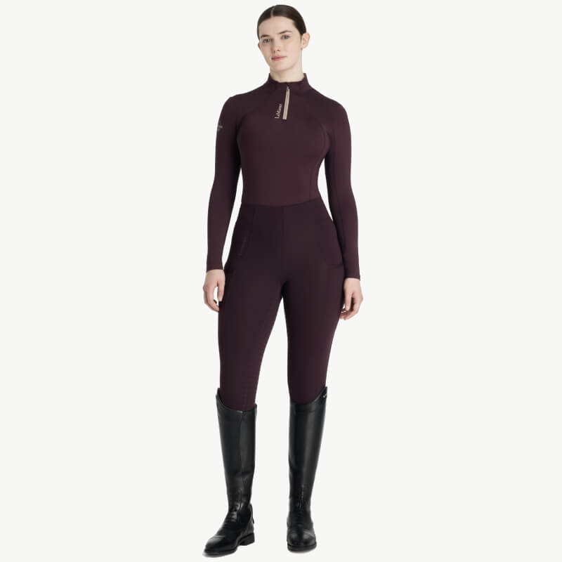 Woman in a LeMieux Classique Baselayer Damson and black boots stands against a white background.