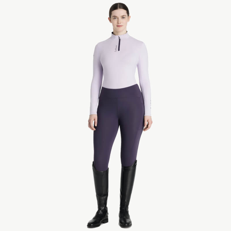 Woman in riding pants, boots, and a LeMieux Classique Baselayer Lilac, standing facing forward.