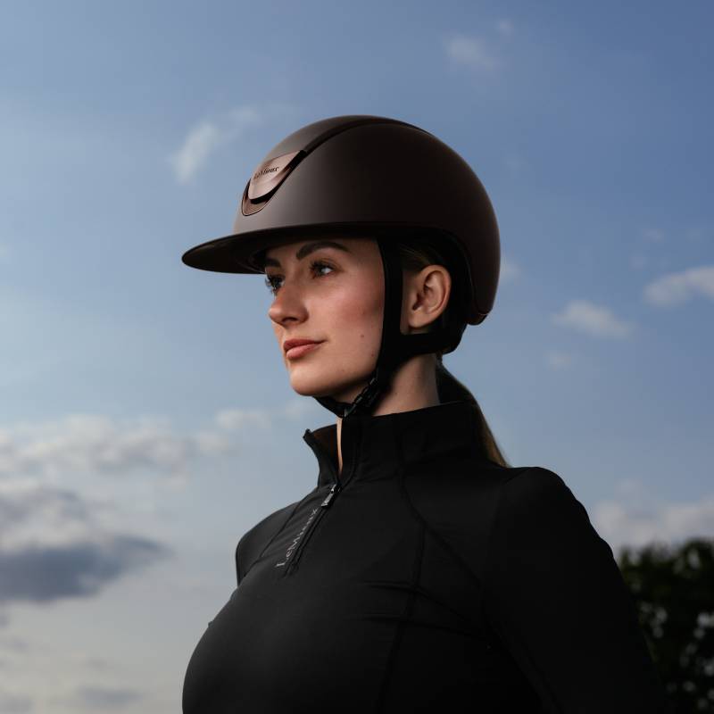 Woman in a LeMieux Verv® Competition Helmet Matte Brown gazes into the distance under blue sky.