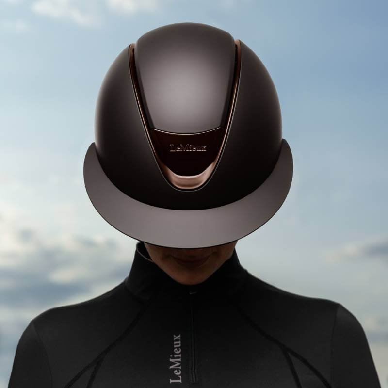Person in a LeMieux Verv® Matte Brown Helmet and black jacket against a cloudy sky background.