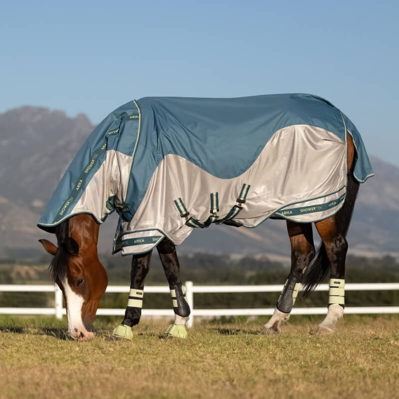 A horse in a LeMieux Arika Camo Shower-Tek Fly Rug Jungle grazes near mountains.