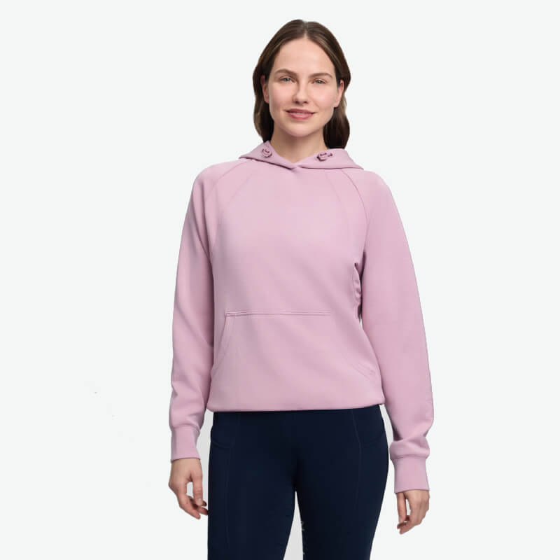 Woman in a LeMieux Nadine Airflow Hoodie Fondant and dark pants smiles against a plain backdrop.