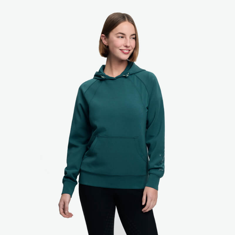 Woman in a LeMieux Nadine Airflow Hoodie Jungle and black pants smiles by a white backdrop.