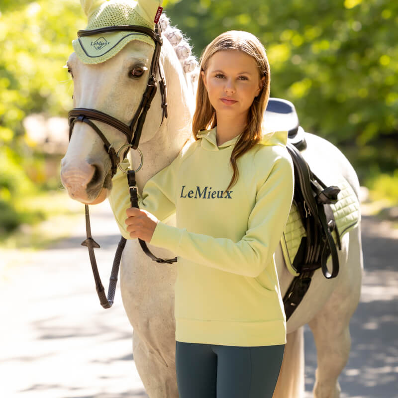 Young woman in LeMieux Young Rider Nancy Hoodie Macaron stands by a white horse outdoors.