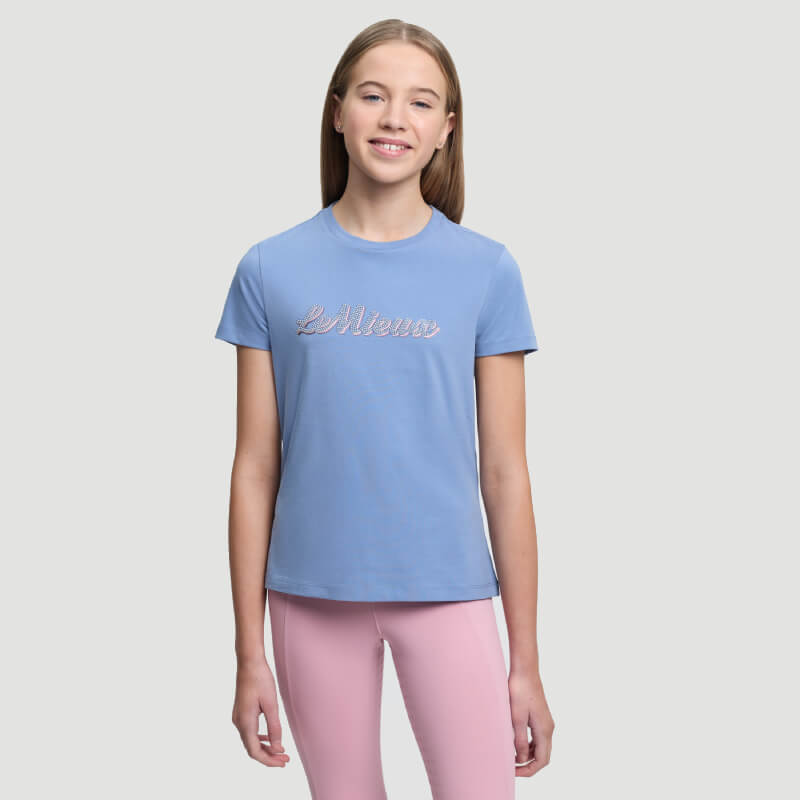 Teen girl in LeMieux Young Rider Arianna T-Shirt Powder Blue and pink pants, smiling at camera.