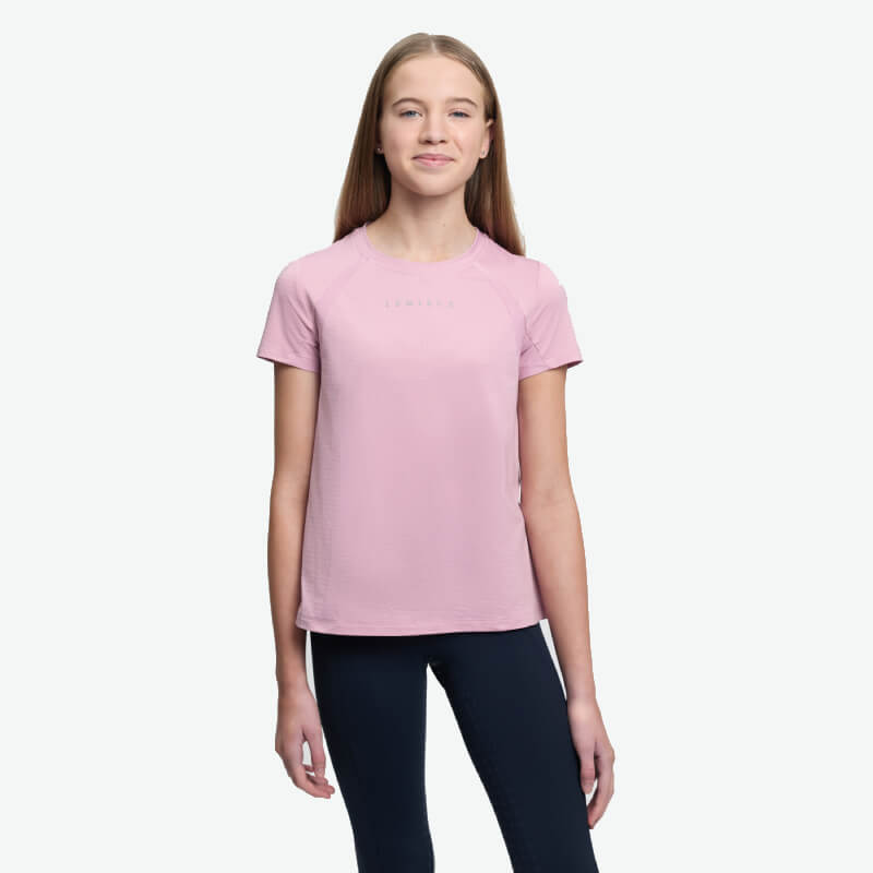 Teen girl in LeMieux Young Rider Sports T-Shirt Fondant and black pants, smiling by a white wall.