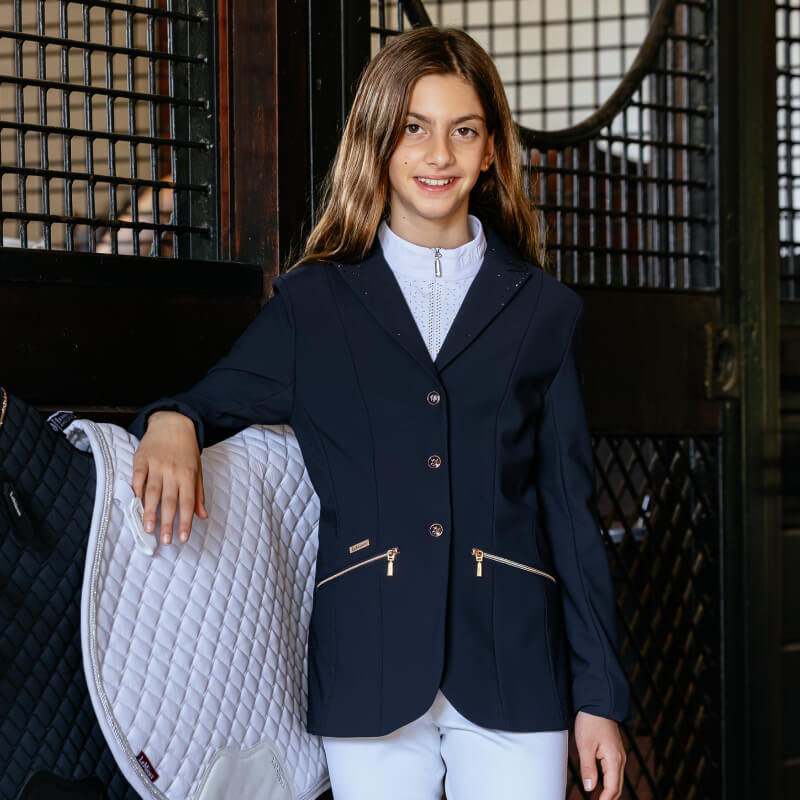 Young rider in LeMieux Brilliance Junior Pro Show Jacket Navy stands by horse stalls.