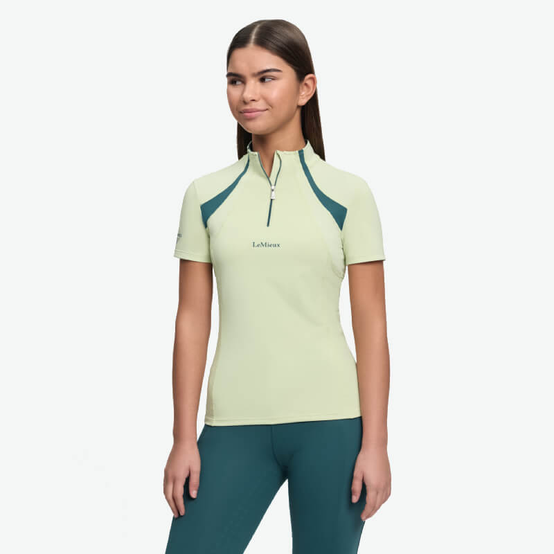 Woman in a light green LeMieux Young Rider Mia Mesh Baselayer Macaron, teal pants, against white.