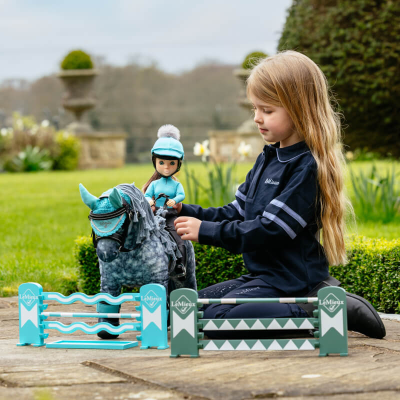 Girl plays outdoors with LeMieux Toy Pony Jump Set Azure/Sage/White and dolls by mini fences.