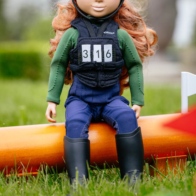 LeMieux Toy Rider in black body protector & number bib sits on an orange pole outdoors.