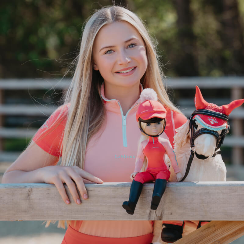 Smiling woman in pink shirt holds a LeMieux Harlow Toy Rider Coral Crush by a wooden fence.
