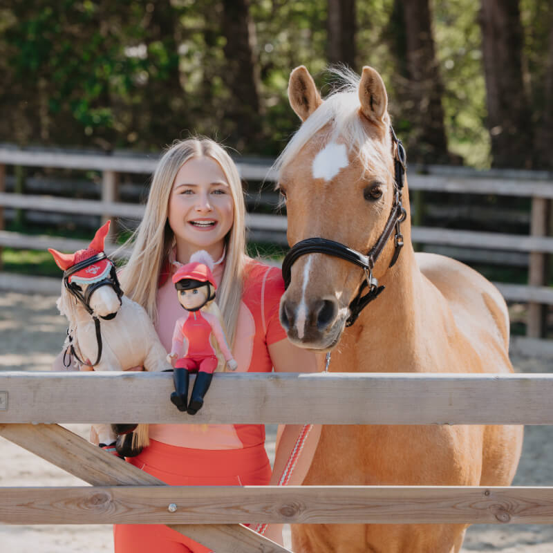 Smiling woman with horse holding a LeMieux Harlow Toy Rider Coral Crush at a wooden fence.