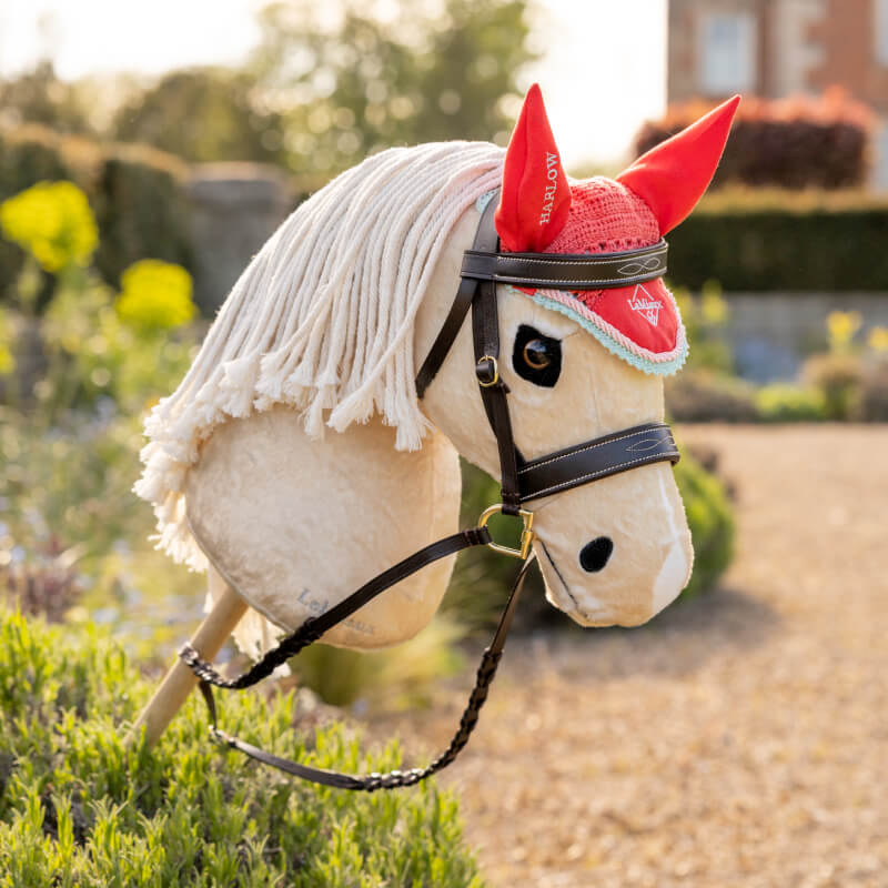 LeMieux Harlow Hobby Horse Fly Hood Coral Crush with red ear covers and yarn mane in a garden.