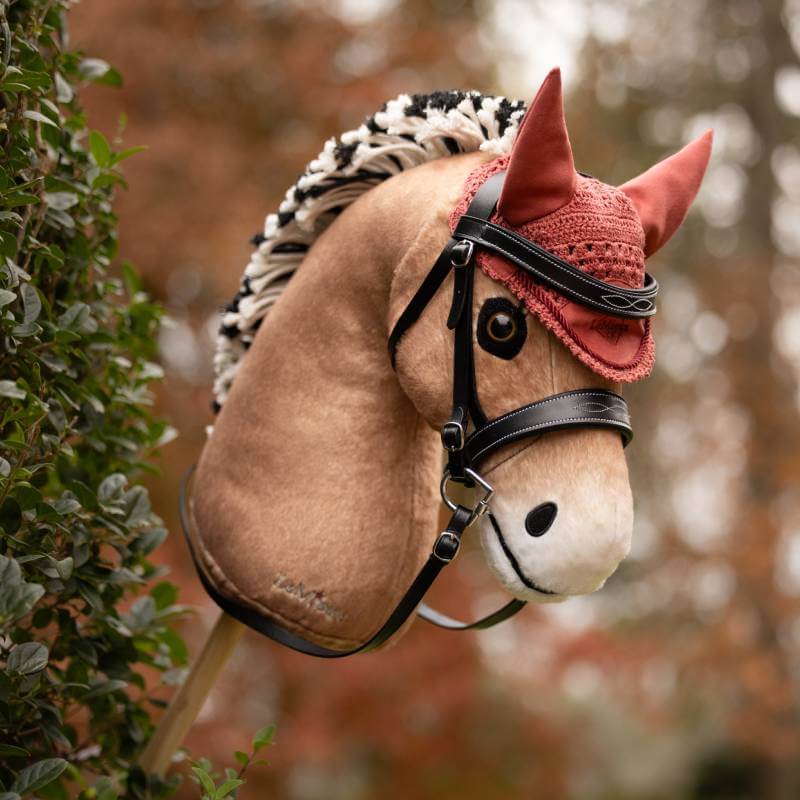 A LeMieux Hobby Horse with bridle and Fly Hood Spiced Earth stands outdoors by greenery.