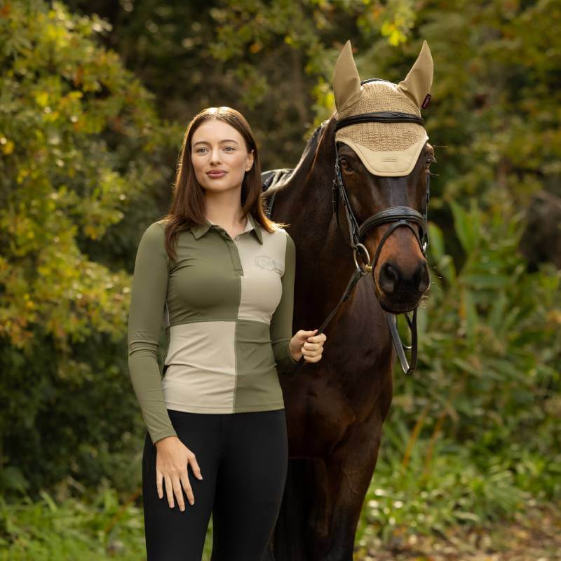 Woman in a LeMieux Harlequin Rugby Shirt Baselayer stands by a dark horse in green scenery.