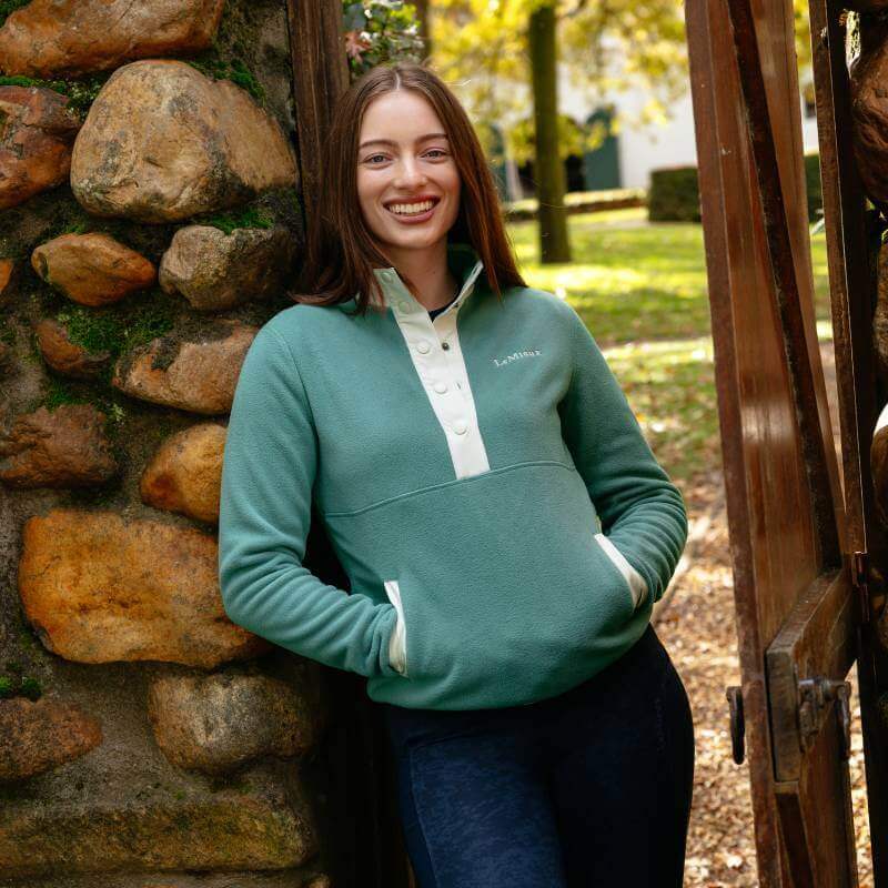 Woman in a LeMieux Piper Popper Fleece Aloe smiles, leaning on a wall near a wooden gate.